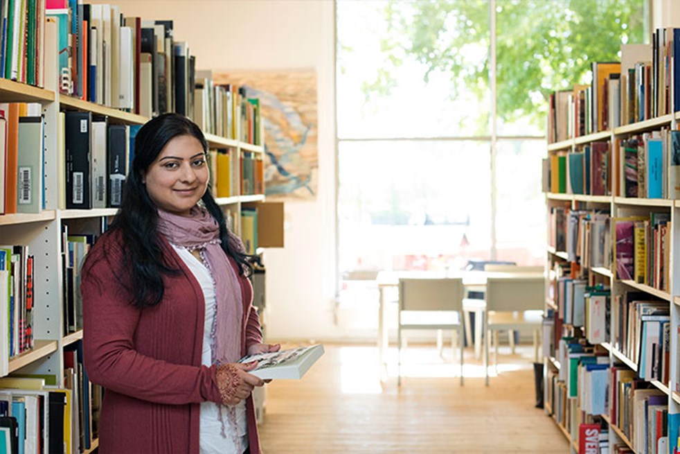 En kvinna med utländskt påbrå  och långt svart hår står mellan två bokhyllor på ett bibliotek.