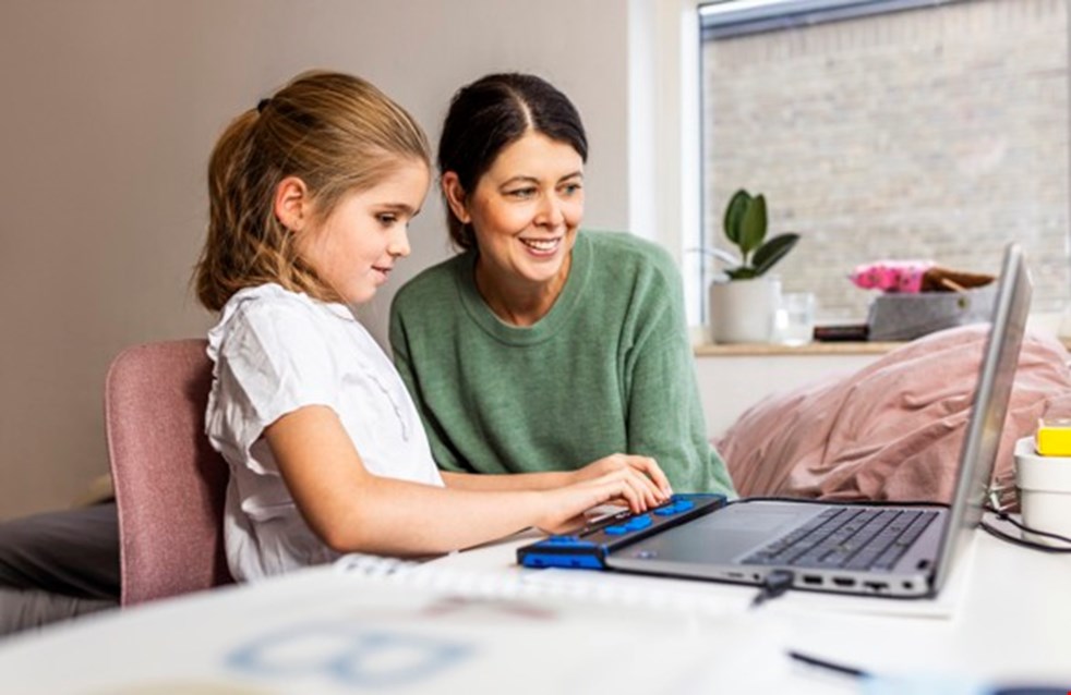 Tioåriga Amanda sitter vid en dator vid ett köksbord. I datorn är en digital punktskriftsdisplay inkopplad. Bredvid flickan sitter hennes mamma. Flickan skriver på sin punktskriftsdisplay och mamman tittar på datorns skärm.