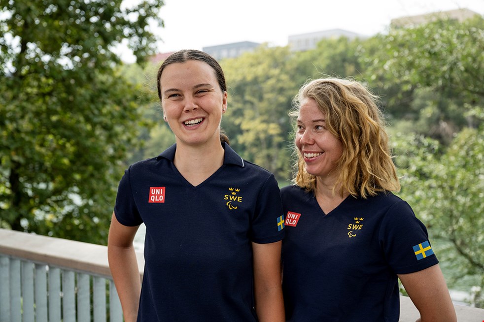 Louise Jannering och Catrin Nilsson står tillsammans utomhus med lummig grönska i bakgrunden. De har mörkblå pikétröjor med den svenska flaggan på och texten "SWE" i gult på bröstet. 