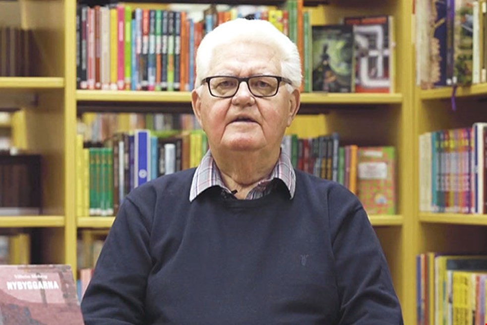 Bild på en äldre man i glasögon framför många bokhyllor fyllda med böcker.