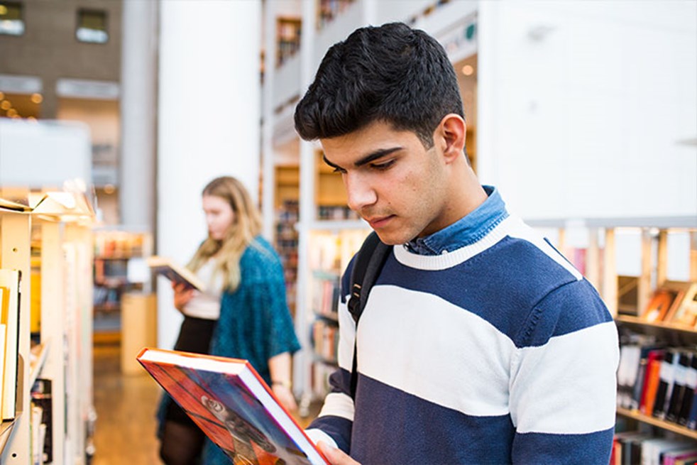 Ung kille står framför en bokhylla på ett bibliotek och läser baksidan på en bok. En ung tjej syns suddigt i bakgrunden.