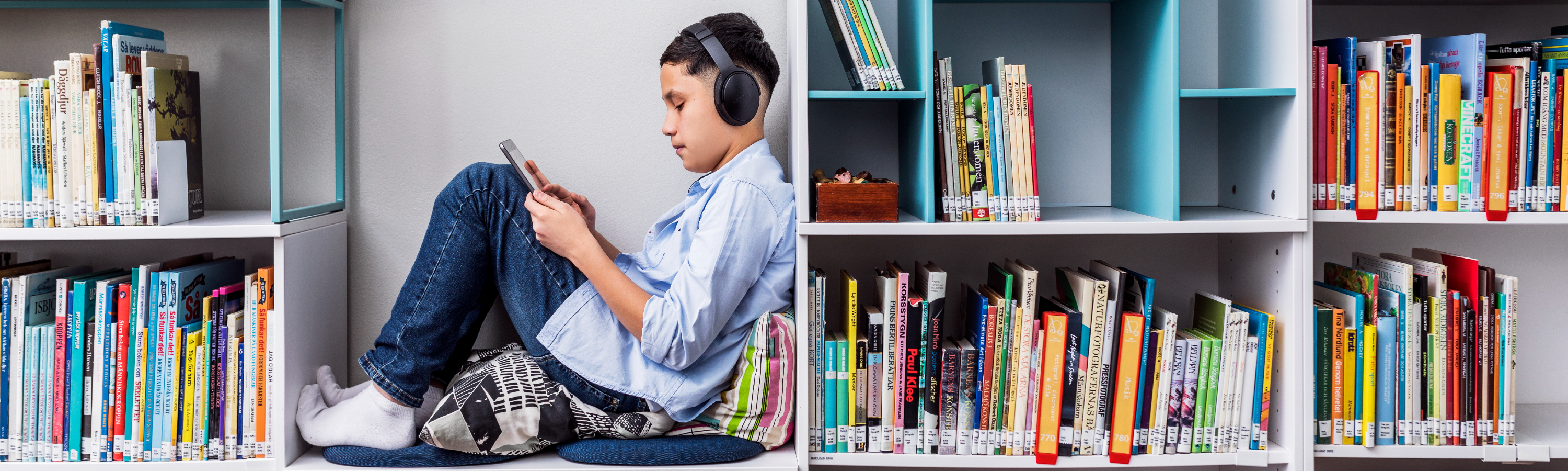 En pojke sitter i en bokhylla omgiven av böcker och läser på en surfplatta. Han har på sig hörlurar.