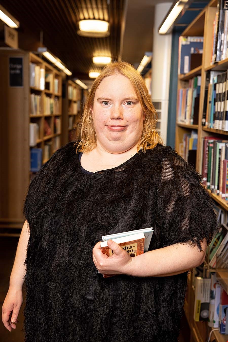 Maila Goman står i halvfigur med ett par böcker i händerna i ett bibliotek.