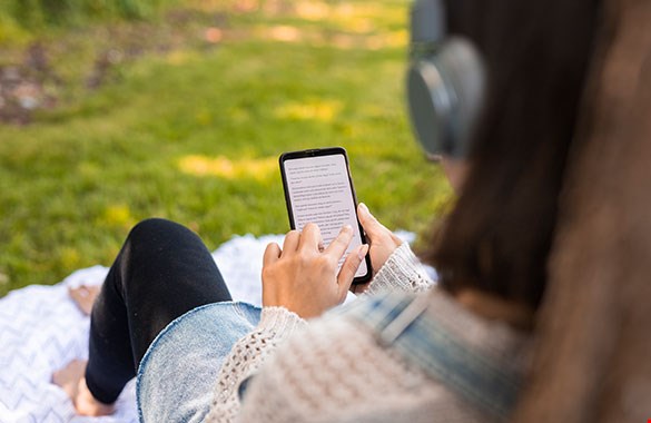 Bild på en ung tjej som sitter utomhus på en filt i gräset med en talbok i telefonen och hörlurar på huvudet. Hon syns bakifrån och man ser inte ansiktet.