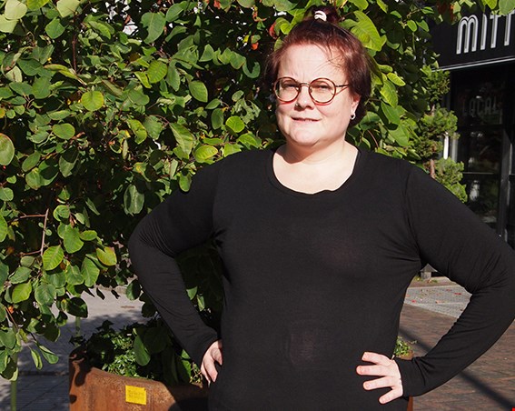 Lina Ydrefelt står utomhus vid ett grönskande buskage. Hon har svarta kläder, glasögon och håret uppsatt i en tofs.