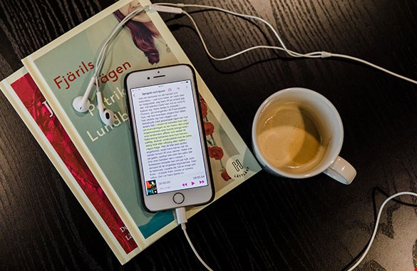 En telefon ligger ovanpå ett par böcker på ett bord. Bredvid står en kaffekopp. Skärmen på telefonen visar en boksida från Legimusappen, en del av texten är markerad i gult. Bilden är tagen snett från sidan.