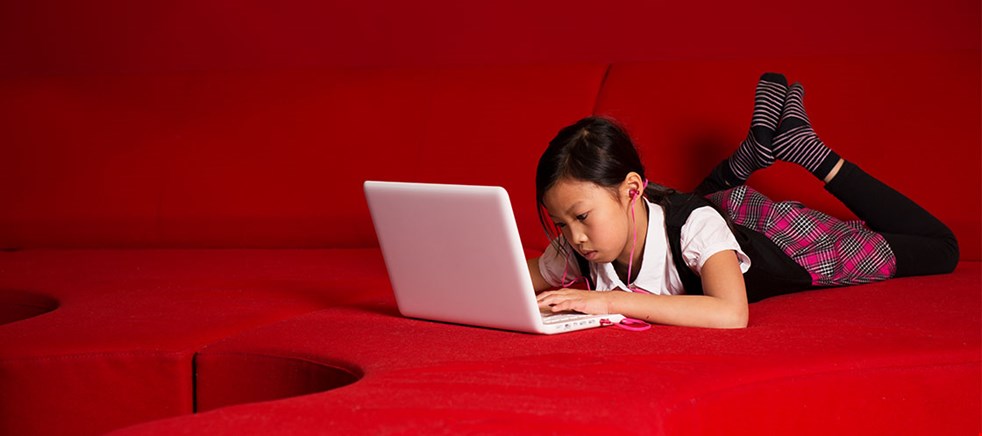 Bilden visar en flicka som ligger på magen i en röd soffa med datorn framför sig och kollar på skärmen.