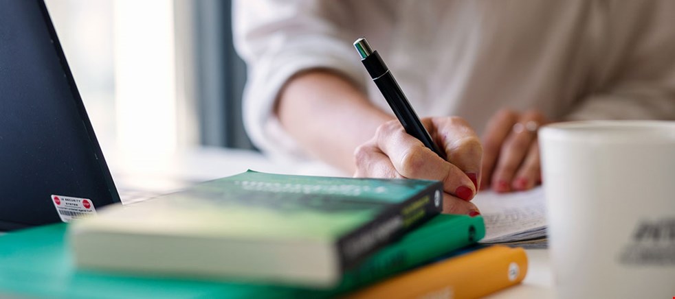 Händer som håller en penna och skriver i ett block. På bordet syns en dator, böcker och en kaffemugg suddigt. Foto: Apelöga