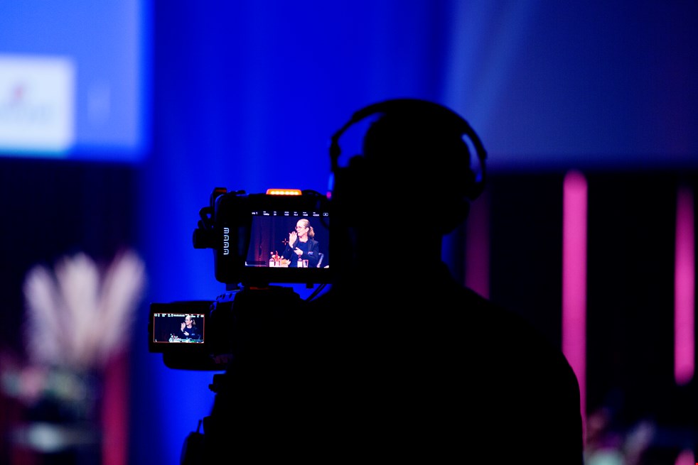 En videokamera filmar en person som sitter på en stor blåaktig scen och tecknar svenskt teckenspråk.
