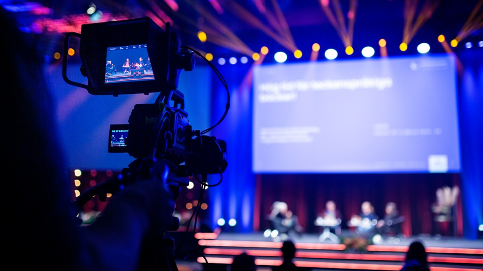 En videokamera är riktad mot en blåaktig scen. I bakgrunden syns en stor digital skärm. På scenen sitter människor som är oskarpa på bilden.