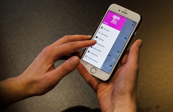 Bilden visar två händer som håller en smarttelefon. På skärmen syns menyn på nya Legimusappen. I bakgrunden syns ett grått golv suddigt.