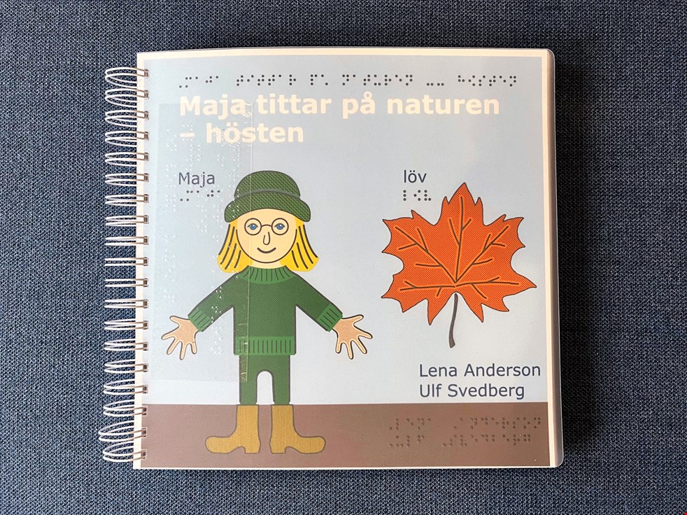 Ett foto av omslaget till den taktila bilderboken Maja tittar på naturen - hösten. Maja har gröna kläder och det ligger ett orange lönnlöv bredvid henne på bilden.
