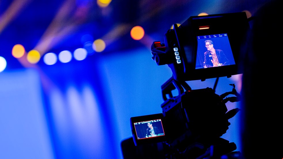 En videokamera står riktad mot en blåaktig studio och i taket finns gula och vita lampor. I kamerans skärm syns en man som ler.