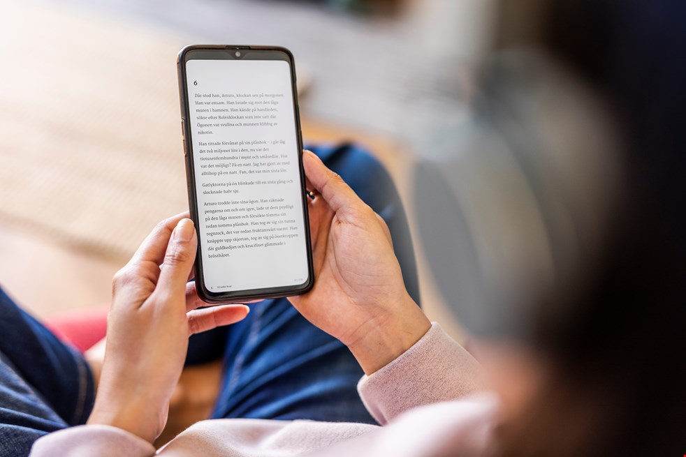 Bilden  visar en närbild på en smarttelefon som hålls av ett par händer av en person som syns med oskärpa bakifrån. På skärmen syns text från en talbok.