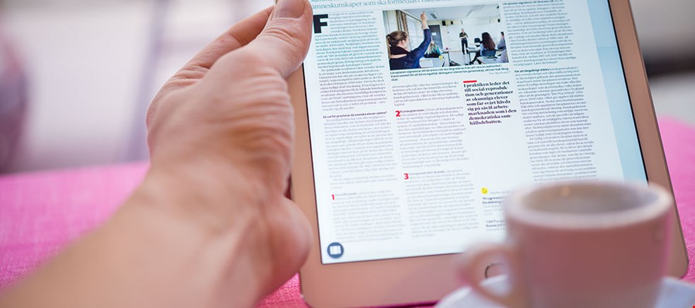 Bilden visar en hand som håller en läsplatta med en dagstidning på skärmen. En kaffakopp syns suddigt i förgrunden.